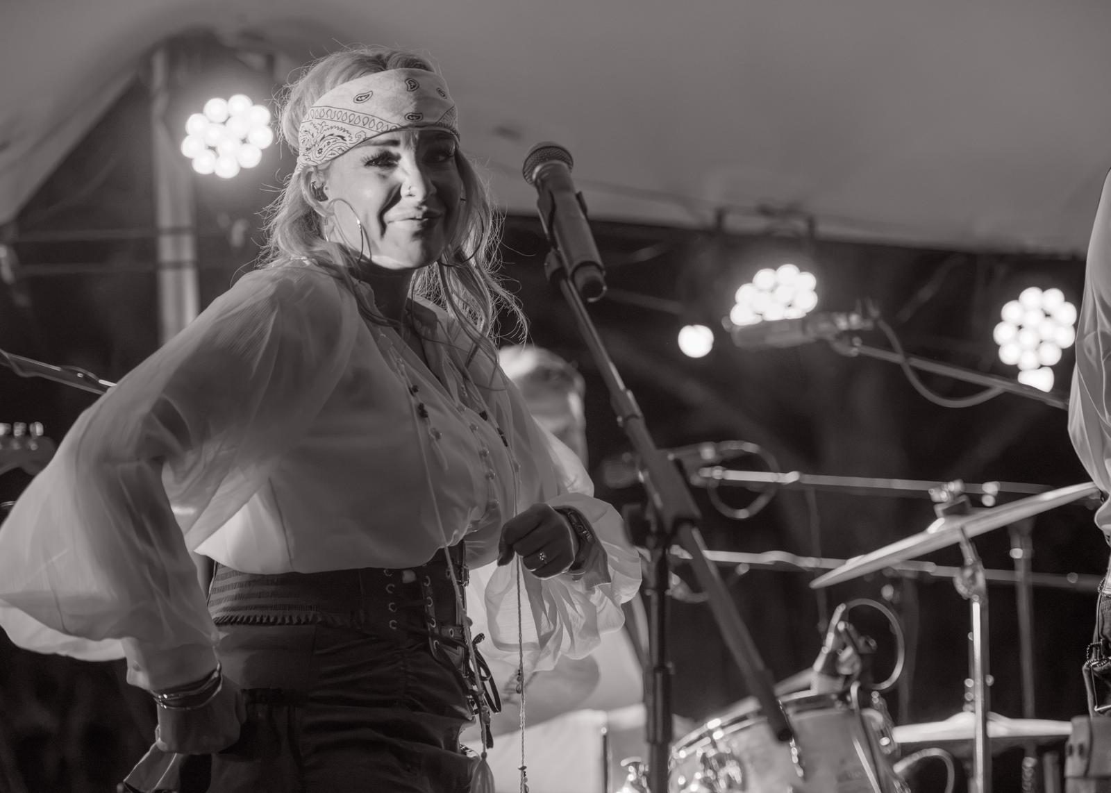 A singer in a white blouse and headscarf smiling at the microphone with stage lights behind